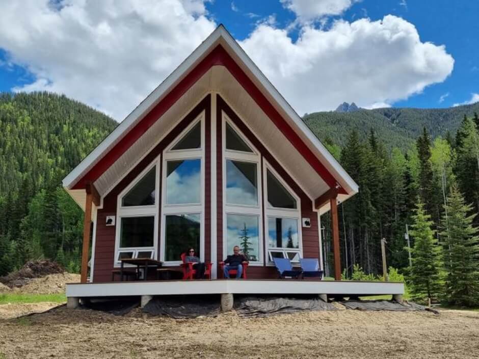 A couple sitting on front deck with the large rake wall window, mountains and trees behind them.